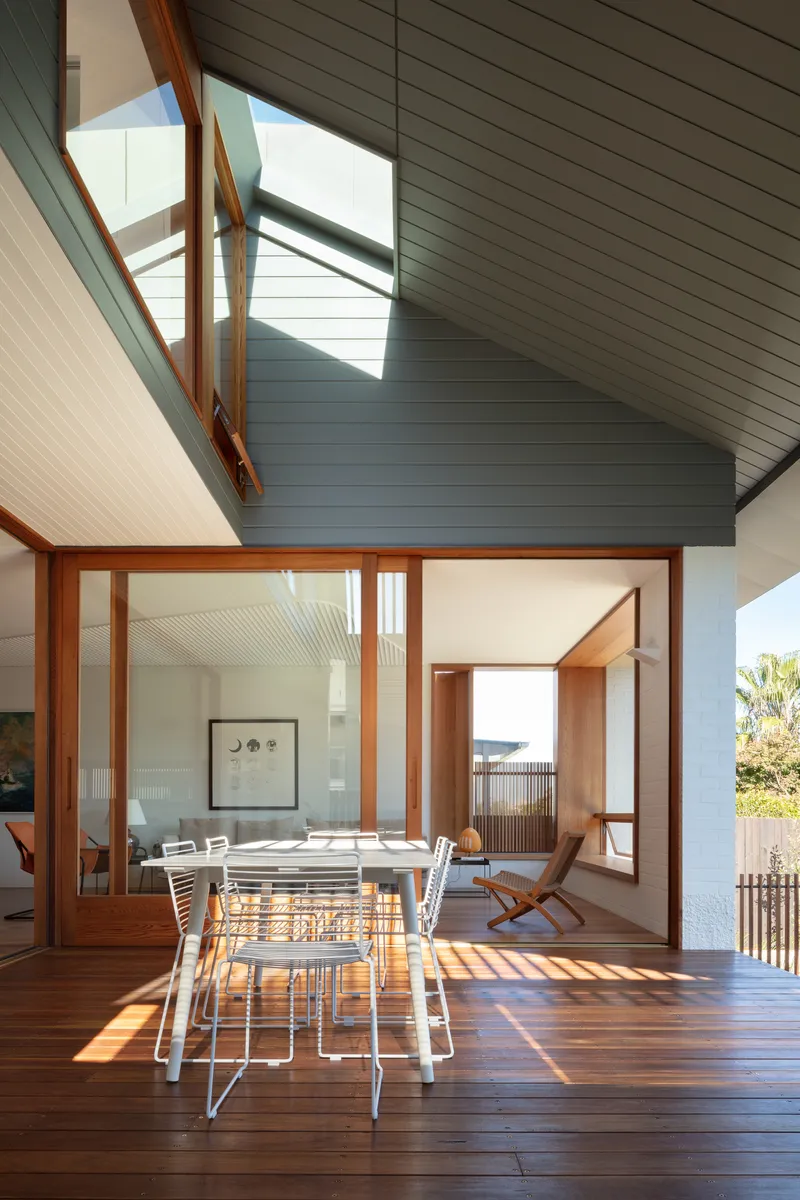 Covered timber deck with dining table framed by glazed walls and angled ceiling above
