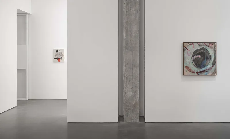 Gallery space with exposed concrete beam dividing white walls hung with framed artworks
