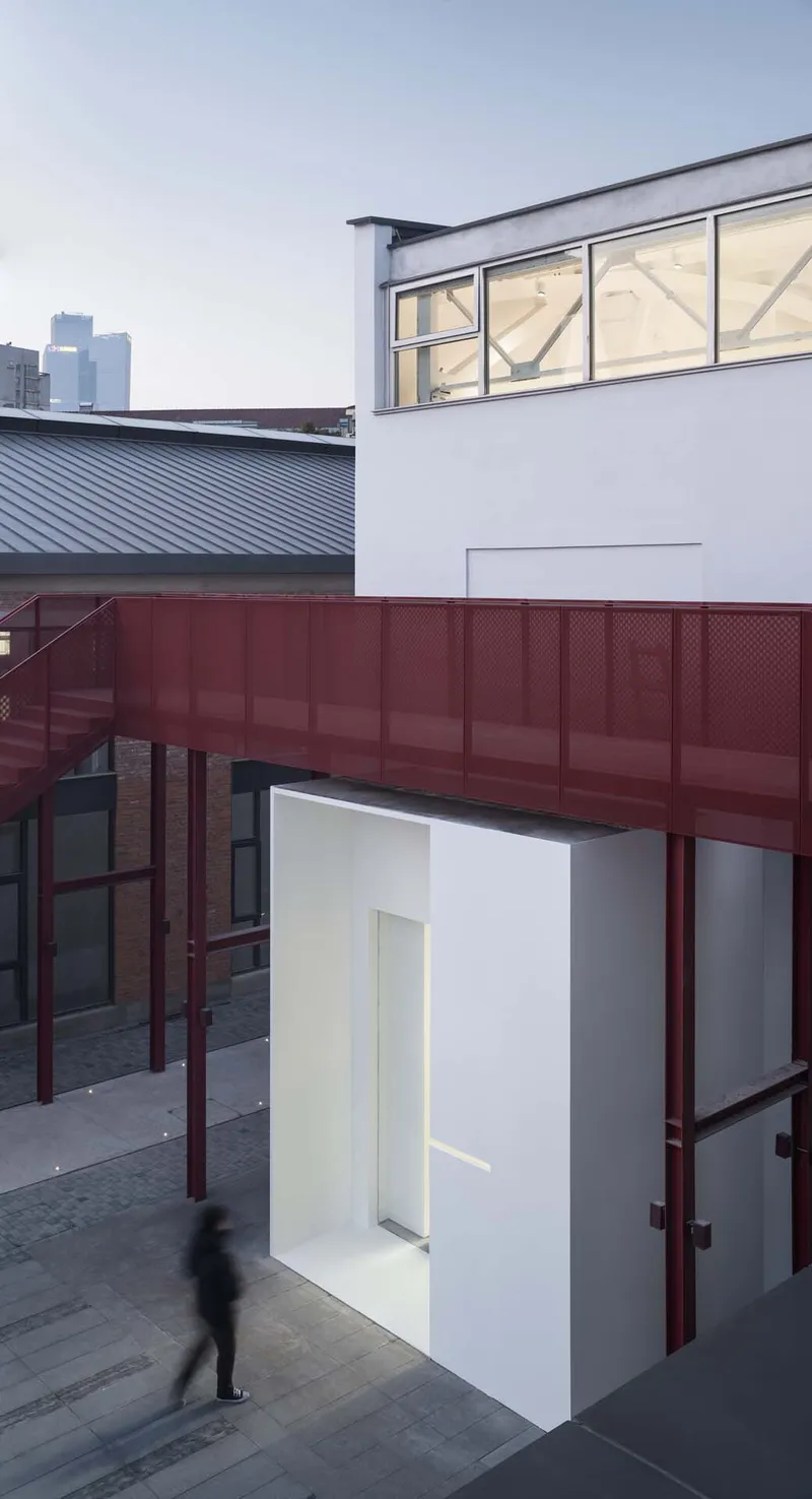 Courtyard view with a red perforated metal walkway over a white cubic volume and a blurred figure