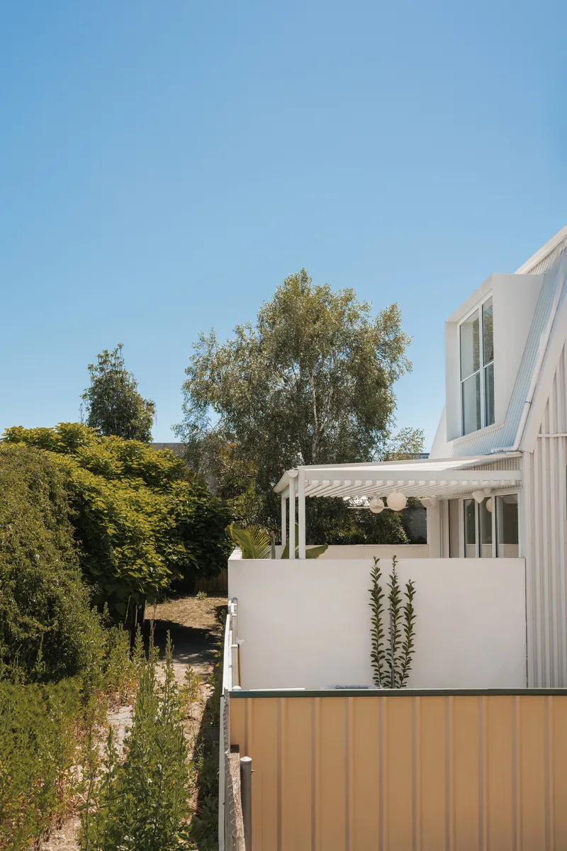 Side elevation showing the white pergola and corrugated metal fence against tall trees in afternoon light