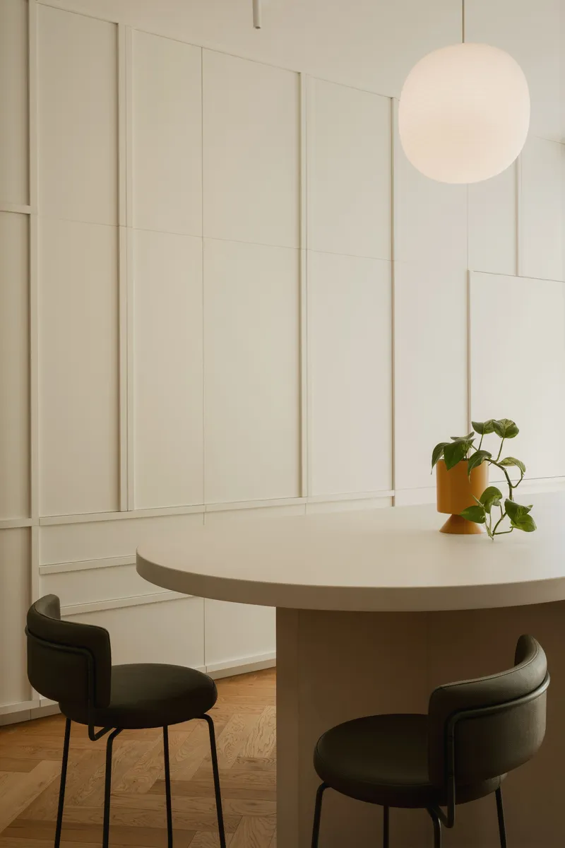 Round dining table with dark upholstered chairs and potted plant under spherical pendant at dusk