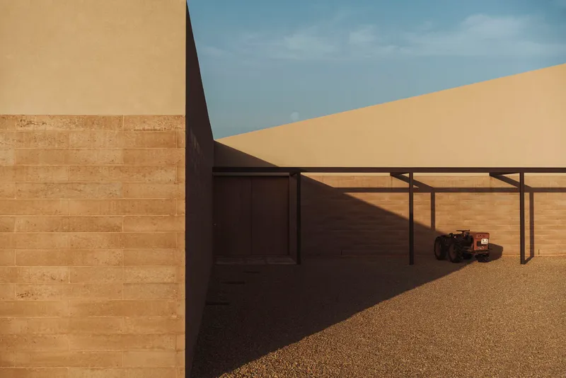Covered exterior walkway with steel pergola casting shadows on gravel courtyard and rammed earth walls