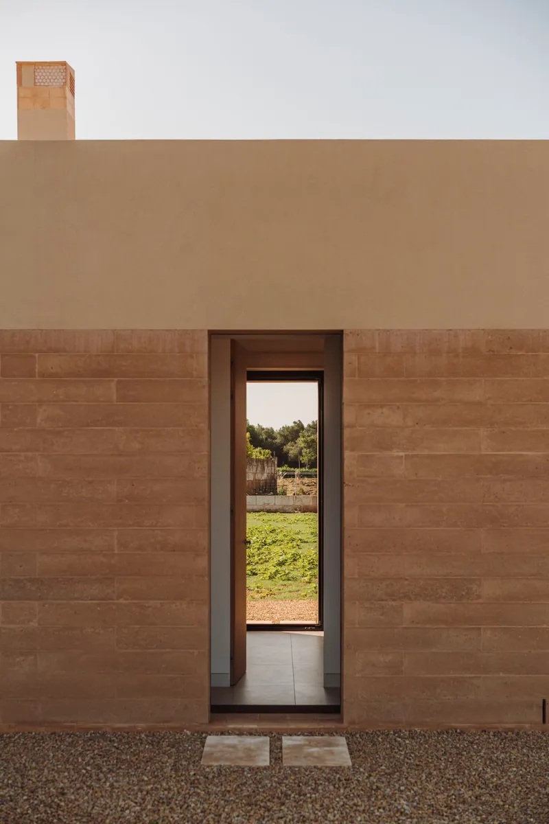 Tall narrow doorway framing view of gravel courtyard and green lawn beyond rammed earth facade
