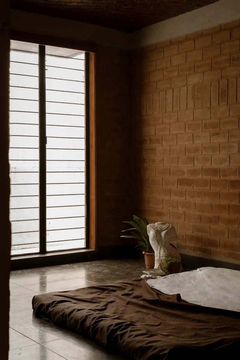 Bedroom with exposed laterite brick wall and horizontal louvered window casting morning shadows across the floor