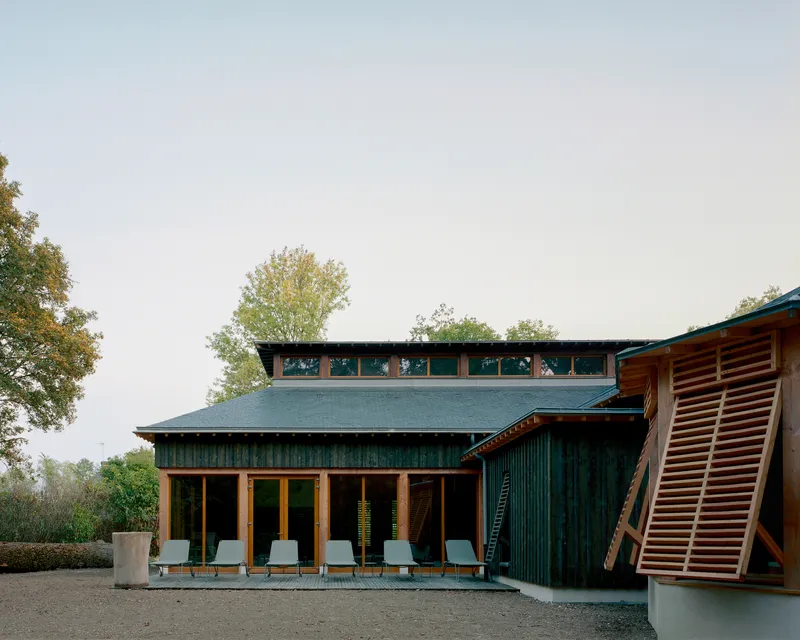 Green corrugated metal facade with timber-framed glazing and a row of chairs on a gravel terrace at dusk
