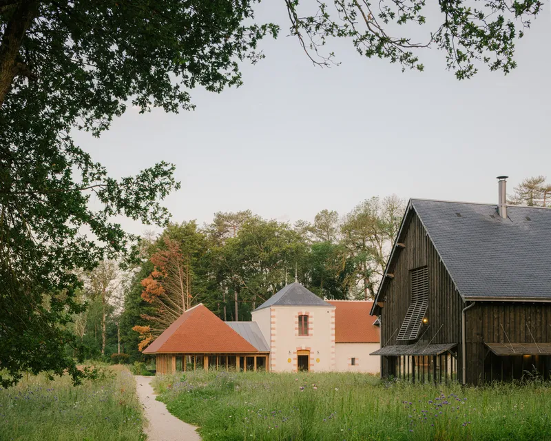 Cluster of buildings with terra-cotta tile roofs and dark timber cladding surrounded by meadow and trees