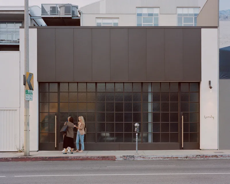 Street facade with gridded glass storefront and dark metal panel upper volume as two pedestrians pause at the entrance