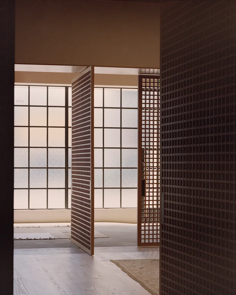 Interior view through open lattice screens toward a backlit translucent grid wall with timber flooring underfoot
