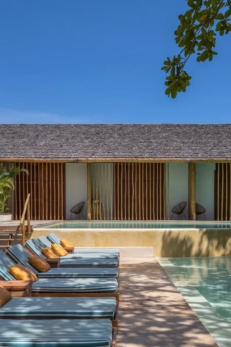 Poolside elevation showing vertical timber slats between stucco walls beneath a grey tiled roof in dappled sunlight