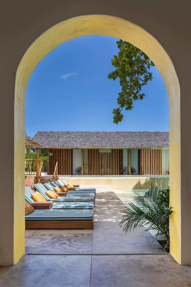 View through a yellow-plastered archway to the pool deck and slatted pavilion beyond with tree branches overhead