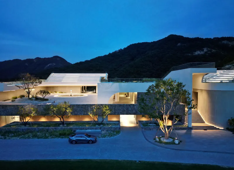 Night view of the white horizontal volumes with ribbed roofs and illuminated openings beneath the dark mountains