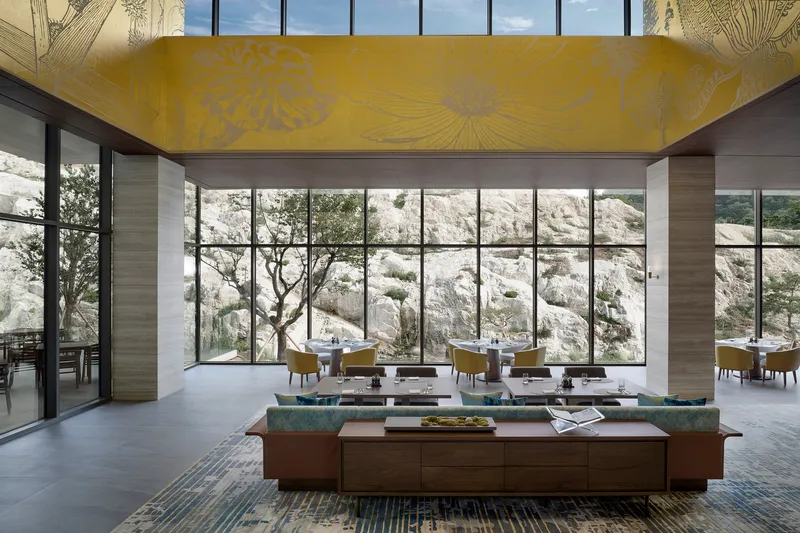 Double-height lounge space with yellow floral ceiling panel and floor-to-ceiling glass overlooking the limestone cliff