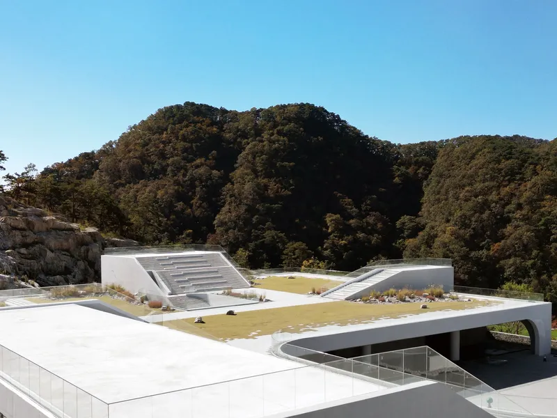 Planted roof terraces with gravel and grass panels step across white concrete volumes against a wooded mountain backdrop