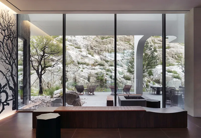 Interior view through floor-to-ceiling glass wall showing terrace with arch and tree against white limestone outcrop
