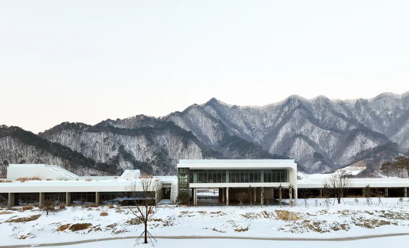 Low horizontal volumes with flat roofs set in a snowy field with bare trees and mountains behind