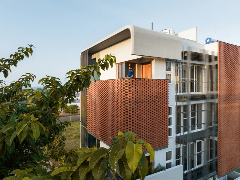 Facade combining perforated terracotta brick screens with vertical metal louvers framed by tropical vegetation