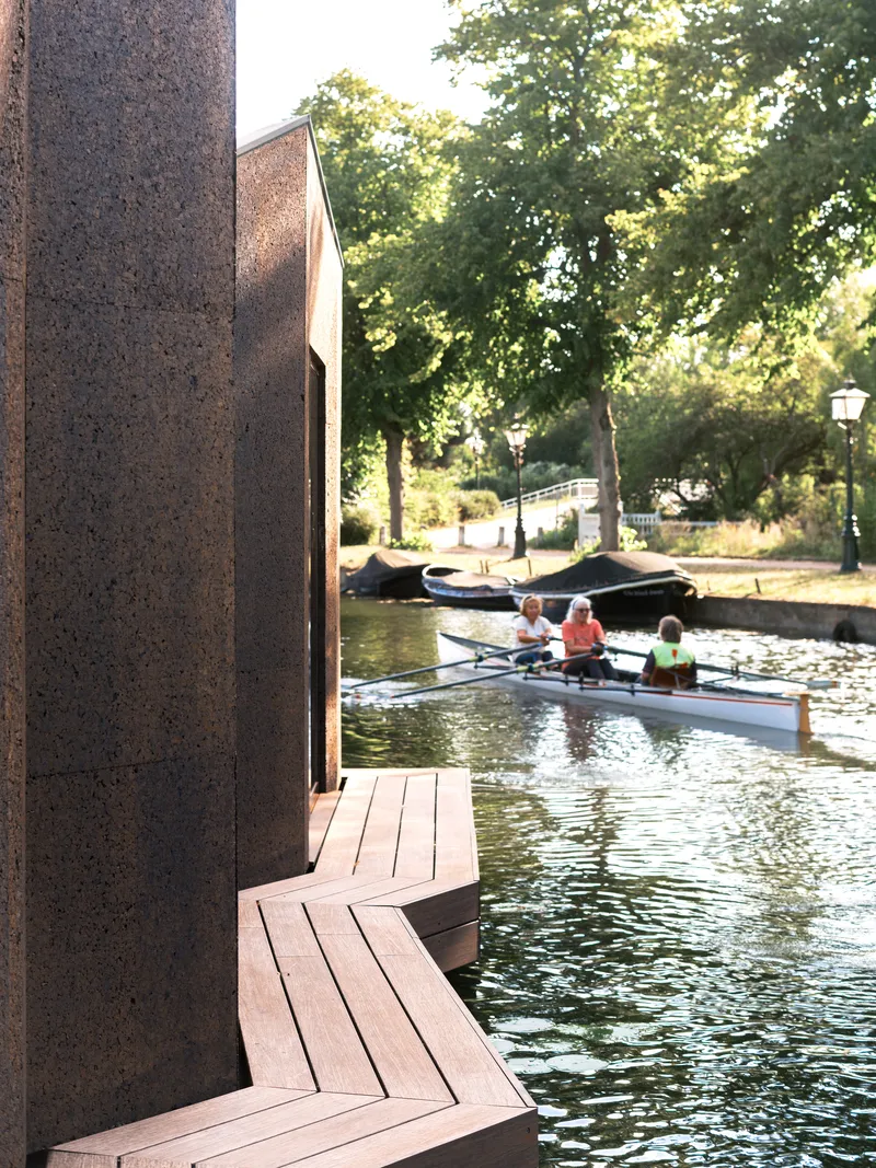 Timber boardwalk wrapping a weathered steel pavilion along a canal with rowers passing by