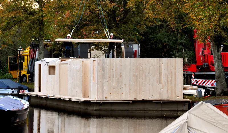 Prefabricated timber-clad floating structure being lifted by crane during assembly on the water