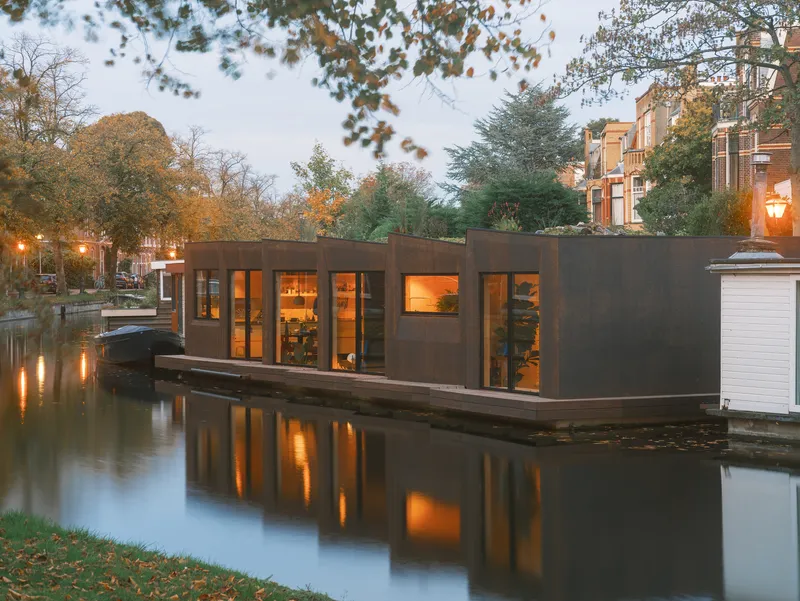 Dark timber-clad houseboat with illuminated interior reflected in the canal at dusk