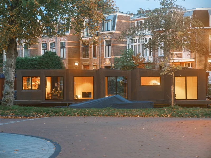 Street view of the houseboat's illuminated glazed facade beneath the brick residential buildings at twilight