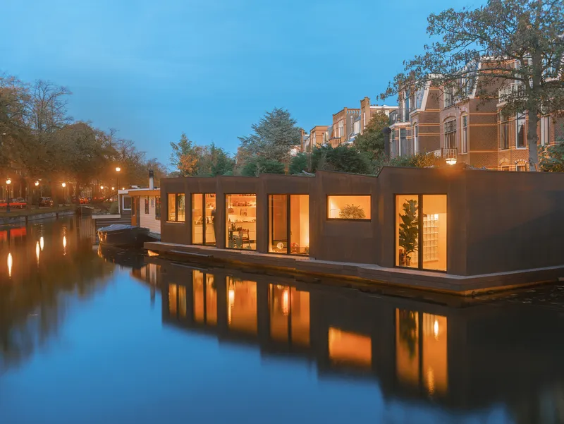Linear facade of the floating dwelling with large glazed openings along the timber deck at twilight