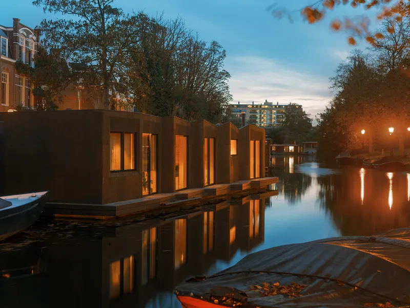 Angled view of the waterside structure with glowing windows and moored boat beside autumn foliage