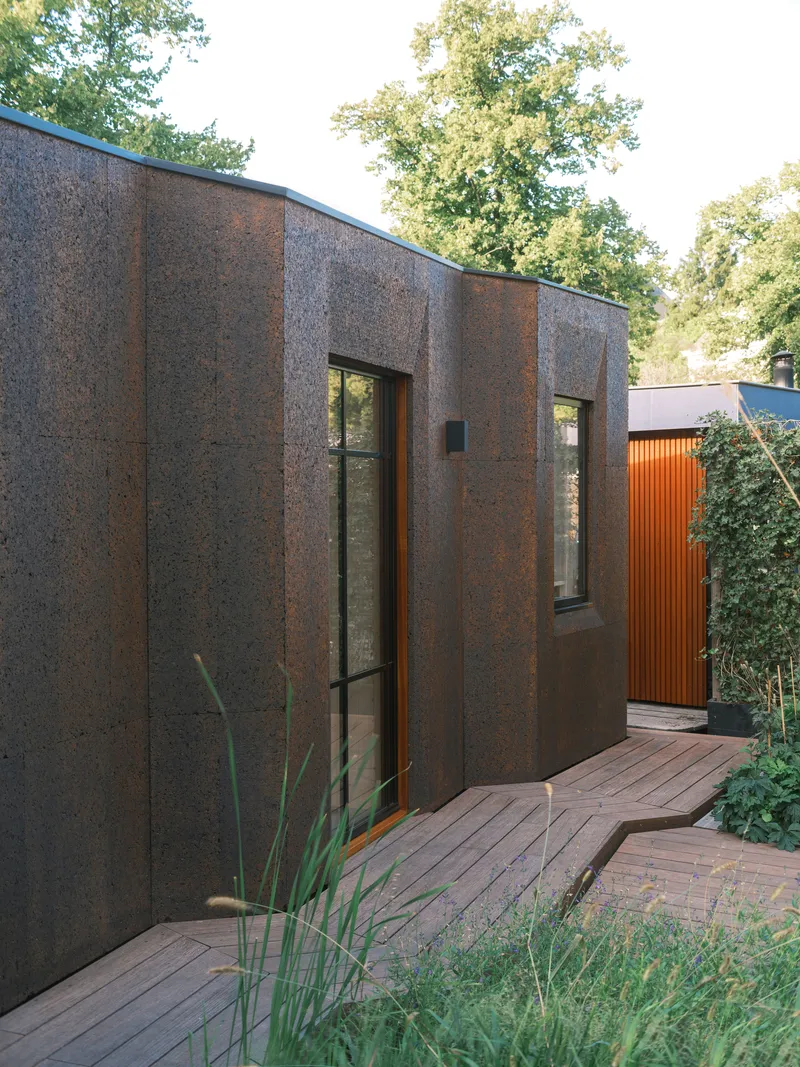 Textured cork panel facade with narrow vertical windows and timber decking in dappled daylight