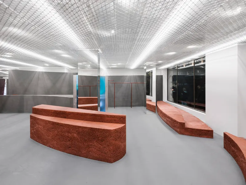 Interior retail space with red textured benches and grey partition walls beneath an undulating metal grid ceiling