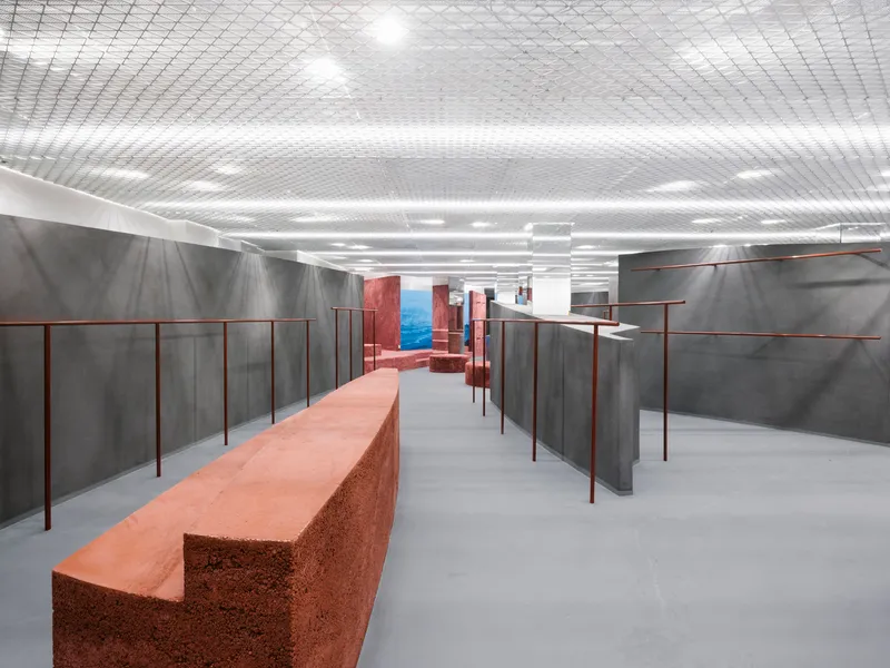 Corridor view between grey partitions with copper rails leading toward red volumes beneath integrated linear lighting