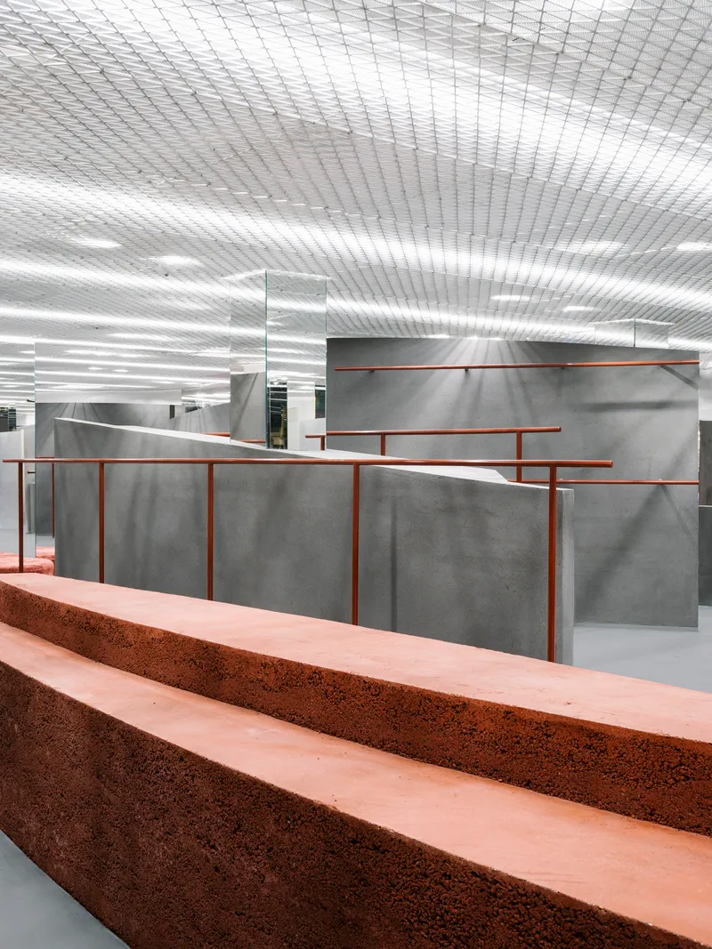 Grey concrete partitions with copper-toned railings beyond a terrazzo bench in the foreground