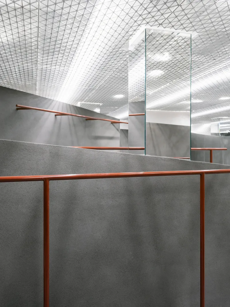 Interior view of grey concrete partition walls with copper handrails beneath a diamond-patterned mesh ceiling