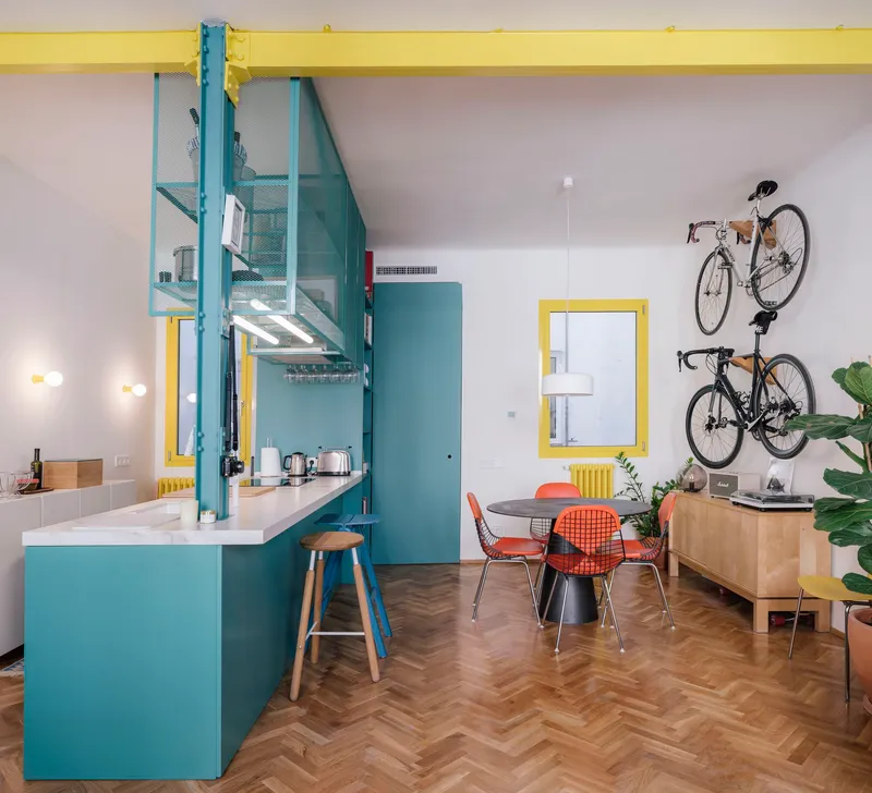 Open living area with teal kitchen island, yellow structural beam, and wall-mounted bicycles above herringbone flooring