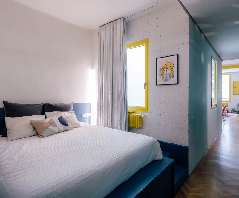 Bedroom with exposed concrete wall, yellow-framed window and yellow radiator beneath sheer curtains
