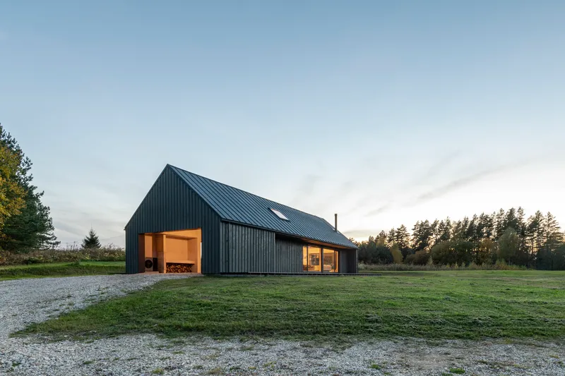 Gabled volume clad in vertical black metal siding with illuminated openings at dusk