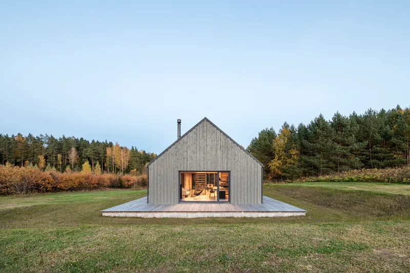 Gabled end facade with full-height glazing and timber deck surrounded by meadow and forest