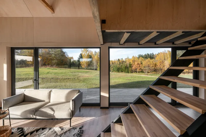 Living area with black steel staircase ascending past floor-to-ceiling glazed doors overlooking meadow