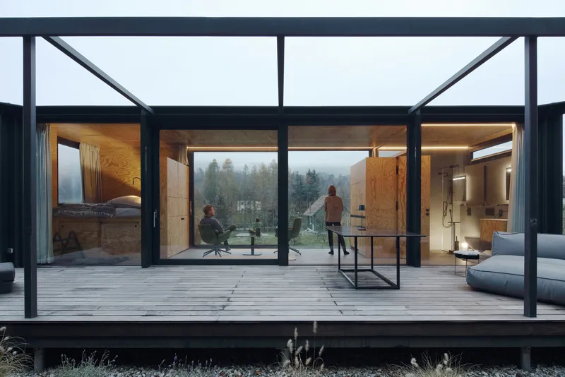 Timber deck terrace beneath steel pergola framing views into plywood-clad rooms with two people in misty weather