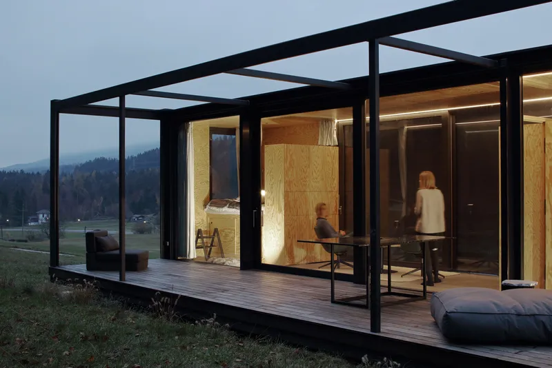 Corner view of the glazed pavilion at twilight showing plywood interior volumes and covered outdoor terrace