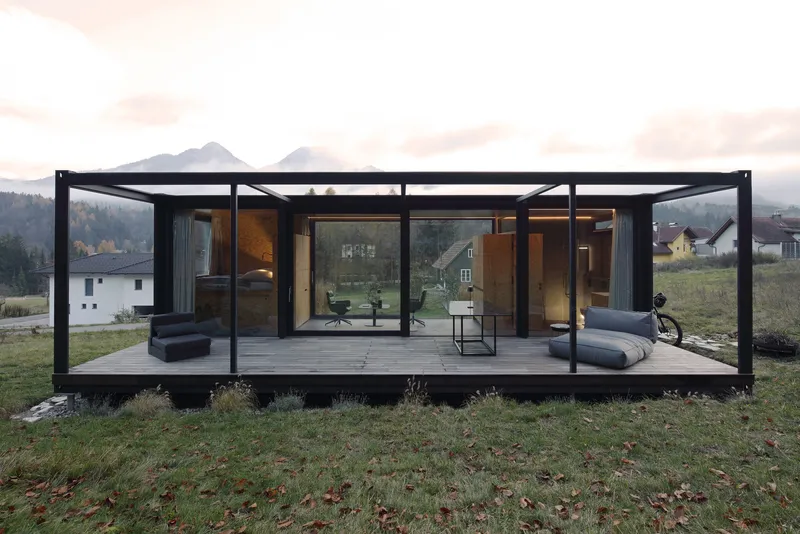 Glass-walled pavilion with black steel frame and covered terrace overlooking a mountain valley at dusk