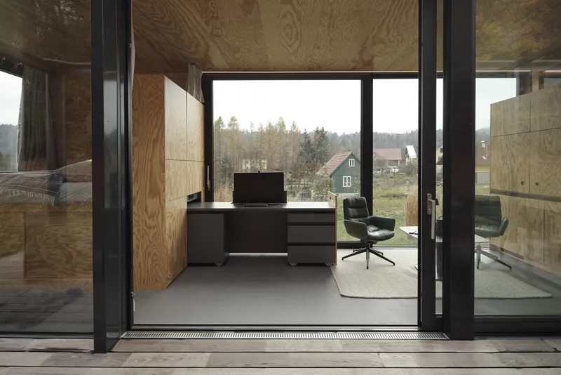 View through open sliding glass doors into a plywood-lined office with desk and chair overlooking distant houses