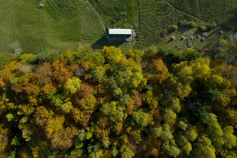 Aerial view of a compact structure nestled at the edge of dense autumn forest and open field