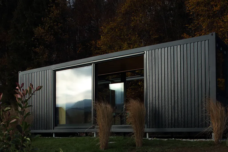 Side elevation showing vertical metal cladding with corner window reflecting the forested hillside