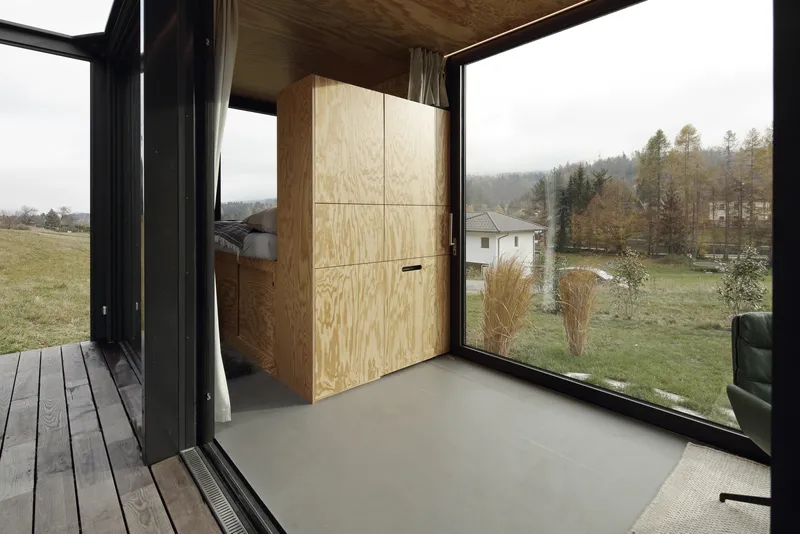 Interior corner with plywood storage wall and floor-to-ceiling glazing opening to autumn landscape