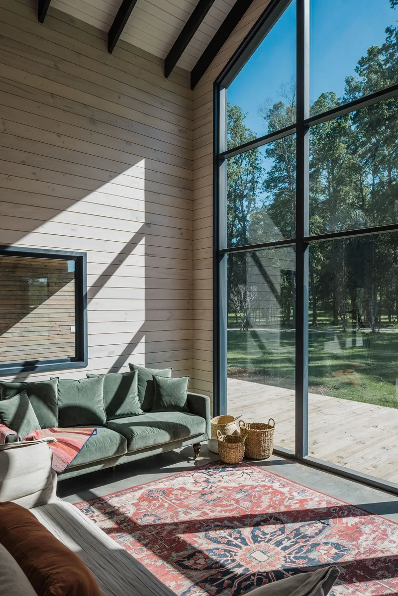 Living space with horizontal wood paneling, exposed rafters and tall black-framed glazing overlooking the meadow