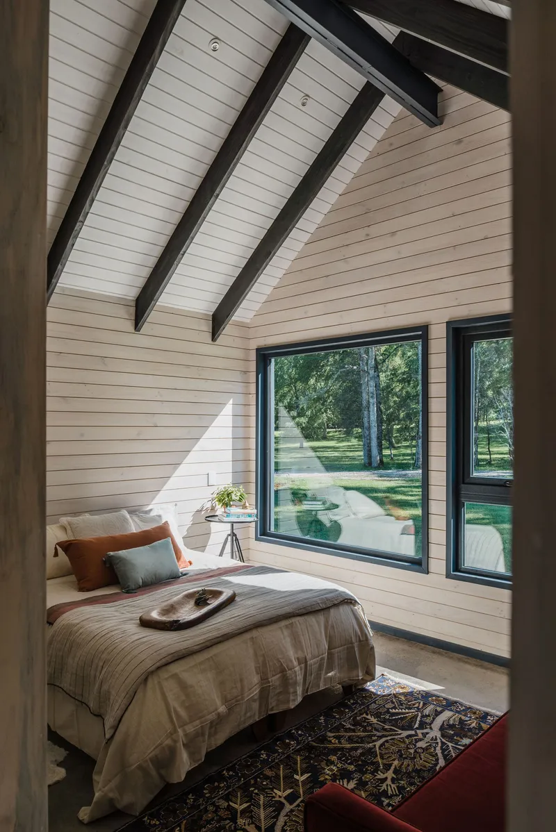 Bedroom with vaulted ceiling, exposed timber beams and floor-to-ceiling windows framing the landscape beyond