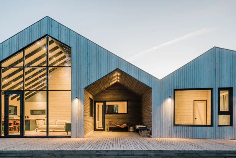 Three gabled volumes clad in corrugated metal with full-height glazing and timber decking at dusk