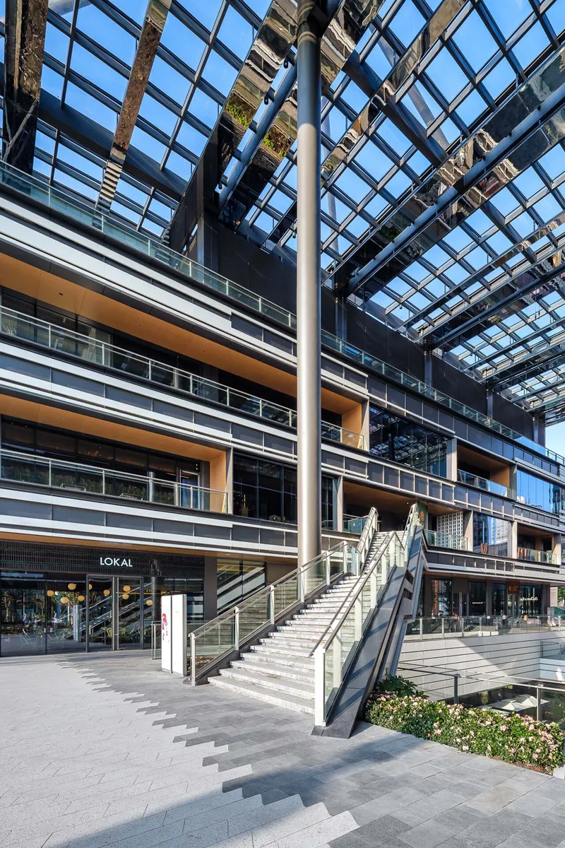 Glass canopy overhead with steel structural grid and terraces below framing an outdoor staircase