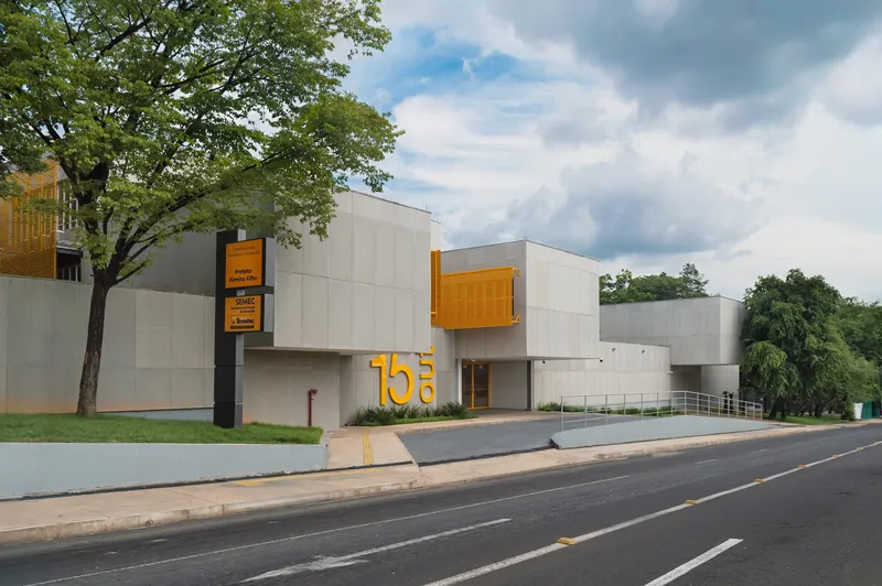 Street facade showing concrete volumes, yellow metal box, and mature trees framing the entrance