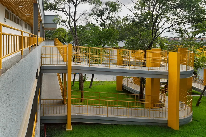 Two-level exterior ramp with yellow steel railings and mesh infill winding around existing trees in a grassy courtyard