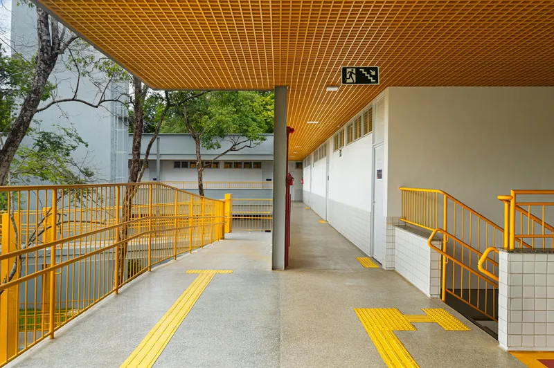 Covered concrete walkway with timber slat ceiling and yellow tactile paving leading toward planted courtyard with mature trees